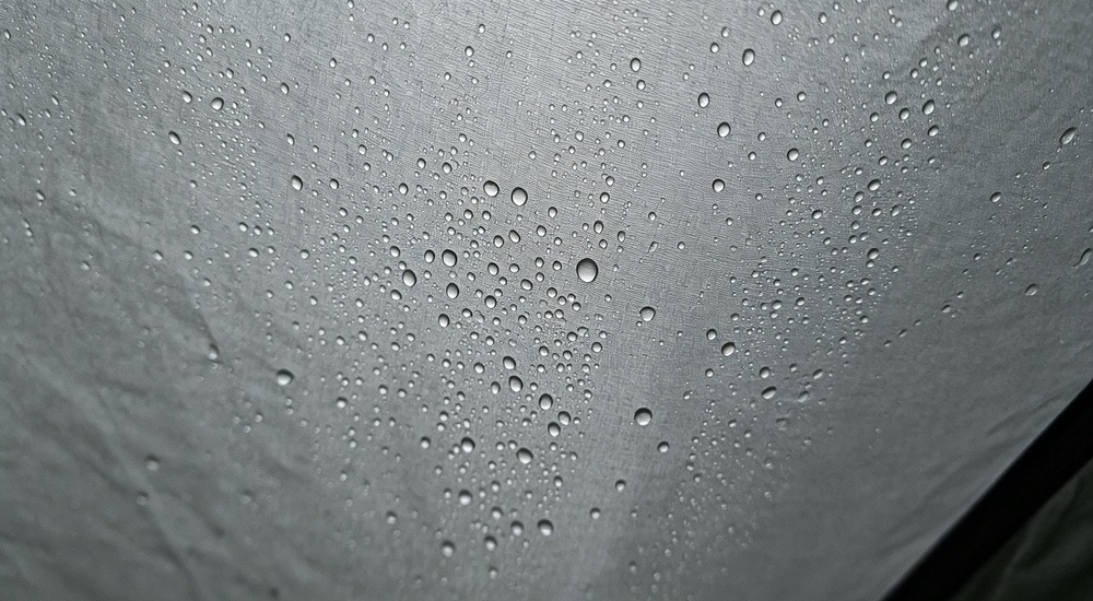 Interior macro photograph showing condensation droplets forming on inner surface of blue tent tarpaulin material, moisture buildup and water drops visible demonstrating humidity and condensation issue in tropical climate shelters