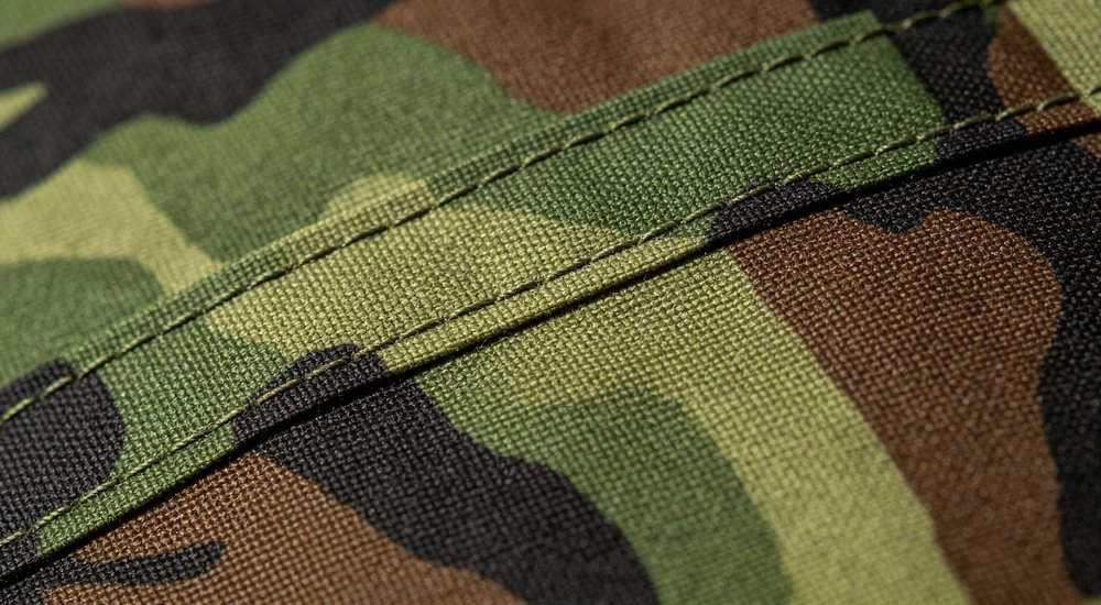 Close-up macro shot of woodland camouflage fabric texture showing green brown black pattern, waterproof Oxford material with detailed reinforced stitching, professional product photography