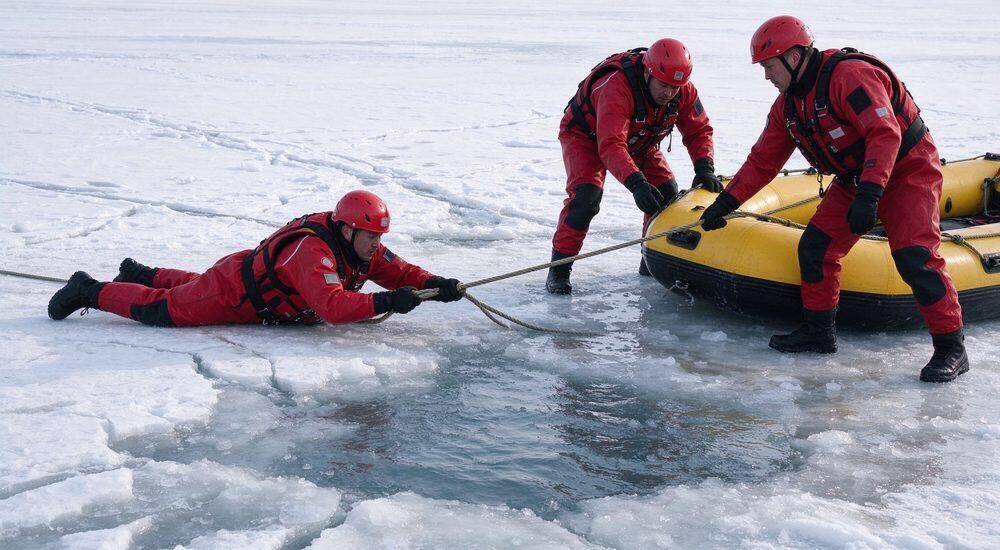 Ice rescue operation on frozen lake with three male rescuers in red drysuits and helmets pulling rescue rope near yellow inflatable rescue raft across cracked ice in cold scenario Ice rescue operation on frozen lake with three male rescuers in red drysuits and helmets pulling rescue rope near yellow inflatable rescue raft across cracked ice in cold scenario